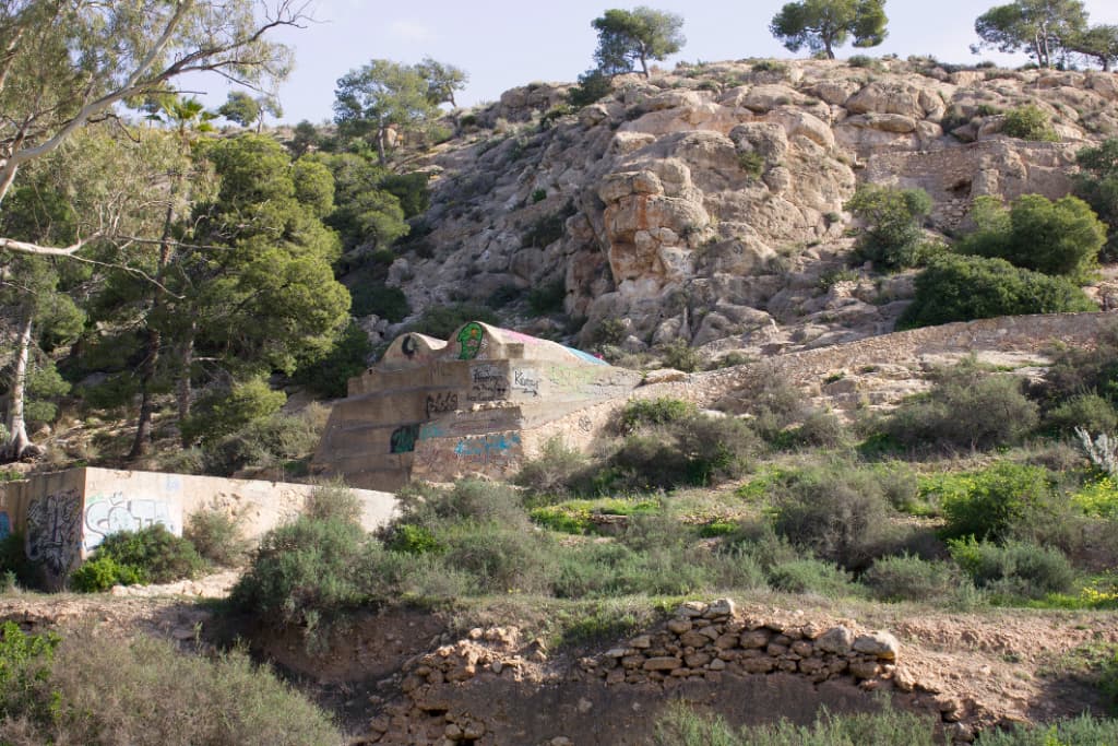 Paisaje de pequeña montaña de roca y vegetación con aljibes y conducciones de agua antiguos en la base de la loma.